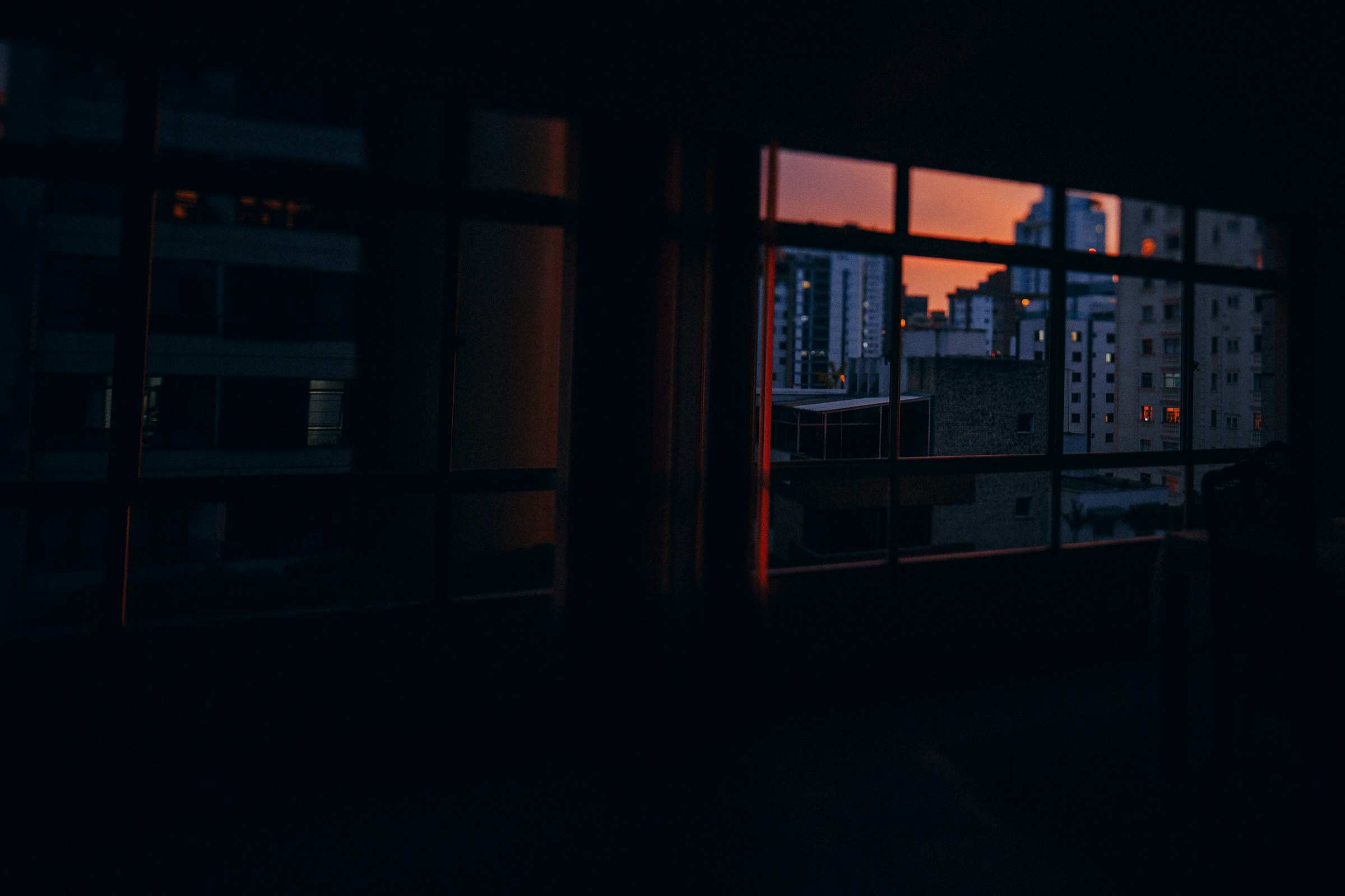 Sala escura com janela para prédios da cidade ao entardecer